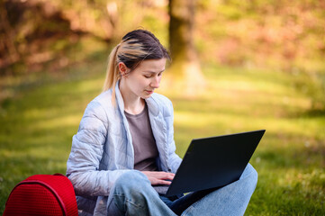 Young girl in spring outwear working on laptop on green grass in parks