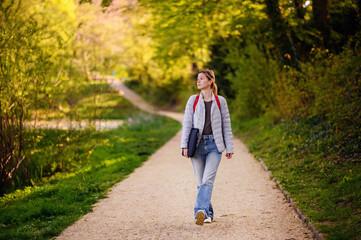Fototapeta premium Young blonde girl in spring outfit walks in green park with red backpack and laptop