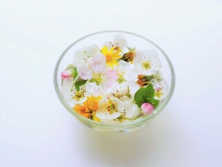 Yellow dandelions, white blooming flowers and green leaves in a glass bowl on a white table