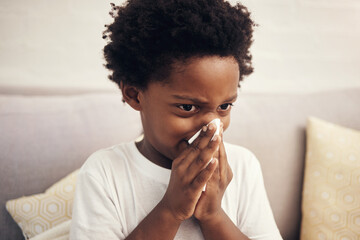 Cold and flu season. Sick african american boy with afro blowing nose into tissue. Child suffering from a runny nose or sneezing, covering his nose while sitting at home. Time to get vaccinated - Powered by Adobe