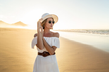 Fashionable woman in white dress and summer hat posing on the sandy beautiful beach