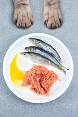 Natural cat food. Fresh raw fish, minced fish, eggs and yoghurt in white bowl with cat paws on grey background Top view