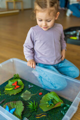 Little girl playing in handmade swamp of green-dyed chia seeds with insect, fish and plant models. Sensory development and experiences, themed activities with children, fine motor skills development