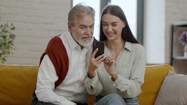 Happy young woman and senior grandfather web surfing in social media on smartphone, watching news online and laughing