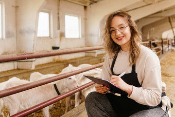 Young smiling woman farmer in apron with tablet on goat farm. Dairy production. Small business...