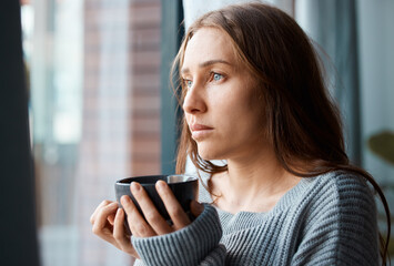 A hot cup of tea to soothe the soul. Shot of a unhappy young woman drinking tea while looking out of the window.