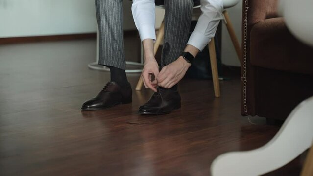 Man in trousers, white shirt and high black socks is pulling and tying laces on his new leather shoes. Businessman is sitting on chair in room next to sofa.