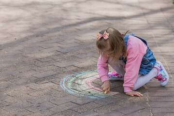 A happy girl in pink clothes draws a rainbow with chalk on the asphalt on the street. Portrait of a little girl drawing with rainbow chalk on a sunny summer day. Creative development of children.