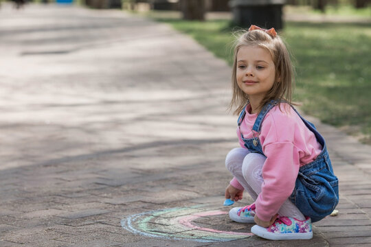 A happy girl in pink clothes draws a rainbow with chalk on the asphalt on the street. Portrait of a little girl drawing with rainbow chalk on a sunny summer day. Creative development of children.