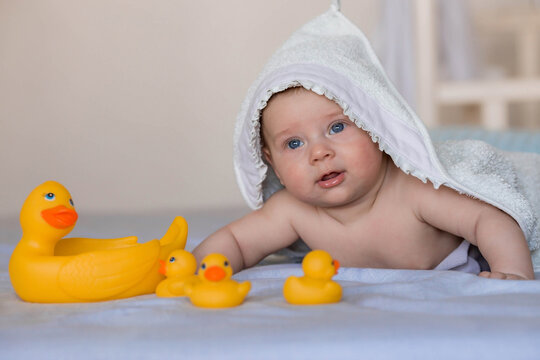 A charming newborn baby is lying on a bed with rubber ducks after bathing. A 4-month-old child with a towel on his head looks at the camera and smiles. Baby bathing and hygiene.