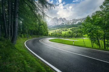 Road in green forest in rainy summer day. Dolomites, Italy. Beautiful mountain roadway, tress, grass, high rocks, blue sky with clouds. Landscape with empty highway through the wood in spring. Travel	