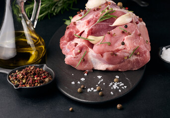 A piece of pork ham on a black board and spices olive oil, salt, rosemary branch and pepper