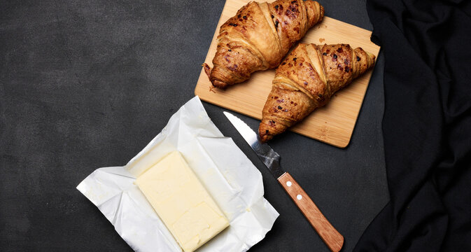 Two baked croissants, a pack of butter and a knife on a black background, top view