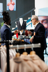 Showroom employee helping stylish client with formal wear, discussing tie fabric in clothing store. Senior man shopping for casual suit, buying fashionable clothes and trendy accessories.