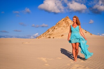 Fototapeta na wymiar Beautiful blonde woman on hot sand with blue sky in the background. Photo taken on the shifting dunes over the Baltic Sea in Leba, Poland. Photo with a shallow depth of field with blurred background.