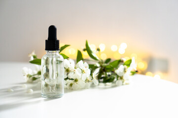 Obraz na płótnie Canvas Clear glass cosmetic dropper bottle and white cherry flowers with bokeh lights, care product mockup