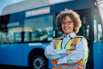 Happy female bus driver with crossed arms at station looking at camera.