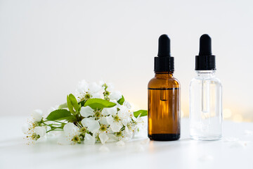 Two clear and amber glass cosmetic dropper bottles and white cherry flowers on white table, care products mockup