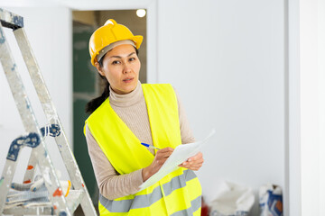 Woman foreman in a protective helmet and yellow vest checks the completed construction work on the drawing
