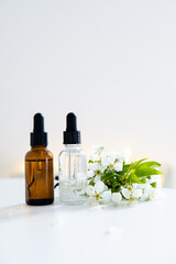 Two clear and amber glass cosmetic dropper bottles and white cherry flowers on white table, care products mockup