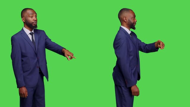 Young businessman waving at someone in studio, telling person that they dropped something over green screen backdrop. Male corporate employee pointing down on floor, greenscreen.
