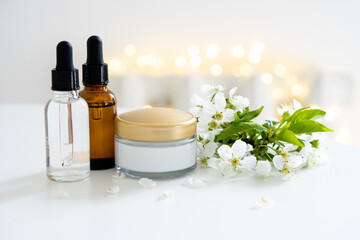 Beauty product dropper bottle and cosmetic cream jar with white cherry flowers on white table background, serum container mockup