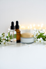 Fototapeta na wymiar Beauty product dropper bottle and cosmetic cream jar with white cherry flowers on white table background, serum container mockup