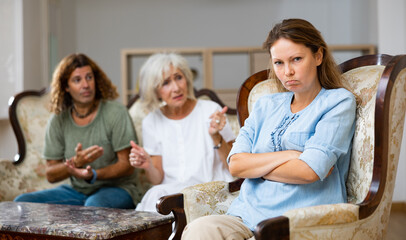 Unhappy casual female sitting on a chair against an upset man and mature woman sitting on the sofa in the living room