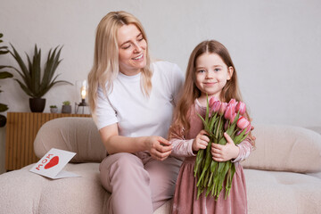 Happy Mother's Day! The child's daughter congratulates her mother and gives her flowers tulips and a card. Mom and girl smile and hug. Family vacation and togetherness.