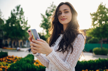 Naklejka premium Portrait of a young fashionable boho woman standing in a city park outside and using phone while looking at the camera.