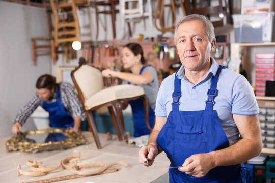 Elderly master restoration worker dressed in overalls poses in working atmosphere of his workshop after working with antique furniture