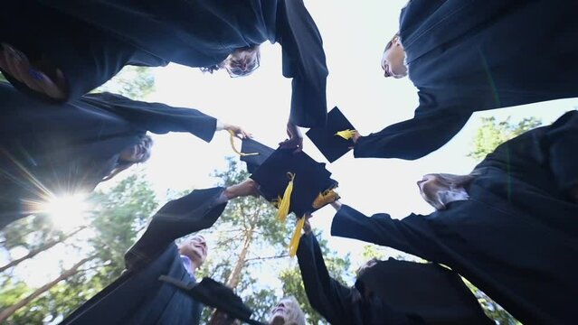 Classmates in graduation gowns toss their hats outdoors. Bottom view. 