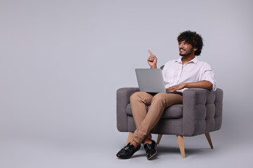 Smiling man with laptop sitting in armchair on light grey background, space for text