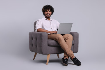 Smiling man with laptop sitting in armchair on light grey background, space for text