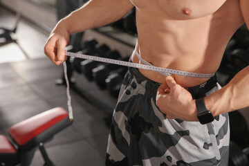 Athletic man measuring waist with tape in gym, closeup