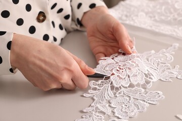 Dressmaker cutting beautiful white lace at table in atelier, closeup - obrazy, fototapety, plakaty