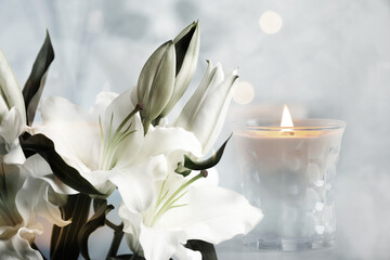 Funeral. Beautiful lilies and burning candle on light blurred background, bokeh effect. - obrazy, fototapety, plakaty