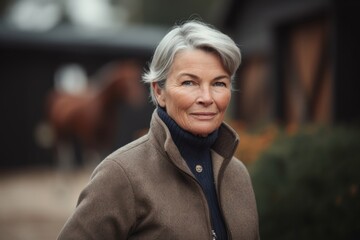 Fototapeta premium Portrait of smiling senior woman in coat standing in front of stable