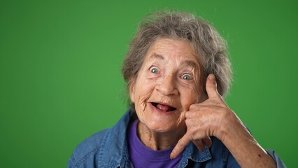Closeup funny portrait of smiling happy elderly senior old woman with wrinkled skin and grey hair smiling and giving call me gesture with hands isolated on green screen background.