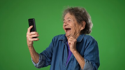 Funny crazy happy elderly old toothless woman taking selfie using mobile cell phone isolated on solid green screen background studio portrait.