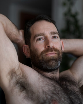 Leaning back shirtless on the sofa, a handsome man expresses contentedness, confidence, and a gentle smile. His thick beard, chest hair, and underarm hair are part of his look.