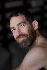 Fototapeta premium Looking over his shoulder while relaxing at home, a man expresses laughter and smiling. He is shirtless, with a thick beard that draws attention to his face.