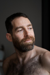 Looking over his shoulder while relaxing at home, a man expresses reflection and contemplation. He is shirtless, with a thick beard that draws attention to his face.