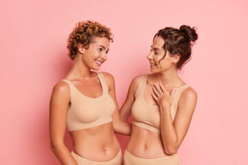 Two well-built girls in beige lingerie pose against a pink wall, dressed in a comfortable top and panties for sports, hugging and laughing, friendship concept, copy space, high quality photo