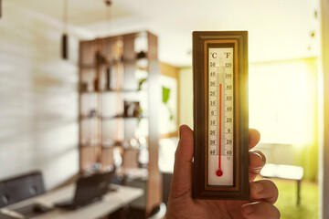 hand with a plastic mercury thermometer shows the temperature in room. Hot sunny weather outside the window. Powerful heating in Russia. - obrazy, fototapety, plakaty