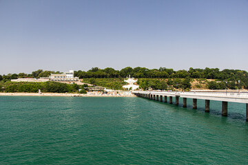 Coastline of a small town with a white pier and green trees. View from the sea