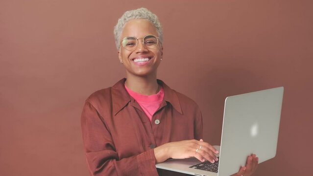 Young happy blonde African American woman typing article in laptop and smiling looking at camera making freelance career or coming up with web resume for new job search stands on brown background.
