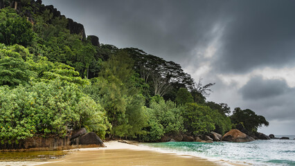 Fototapeta na wymiar Rainy day on a tropical island. The waves of the turquoise ocean spread over the wet sand of the beach. Lush tropical vegetation on the hill, boulders in the water. Cloudy. Seychelles. Mahe.