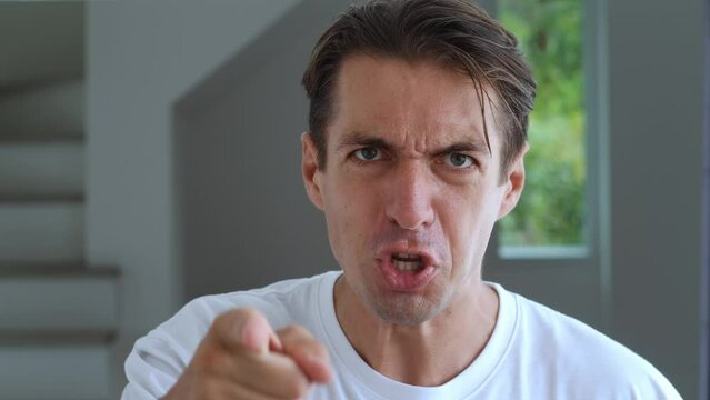 Annoyed and aggressive man screams, talks to camera, curses and quarrels in rage. Yelling annoyed person insults opponent and threatens with pointing finger at home interior indoors. Domestic violence