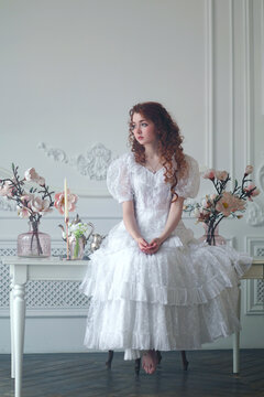 Young red haired woman in white vintage dress sitting on table decorated with flowers against white wall. Historical portrait of female adult